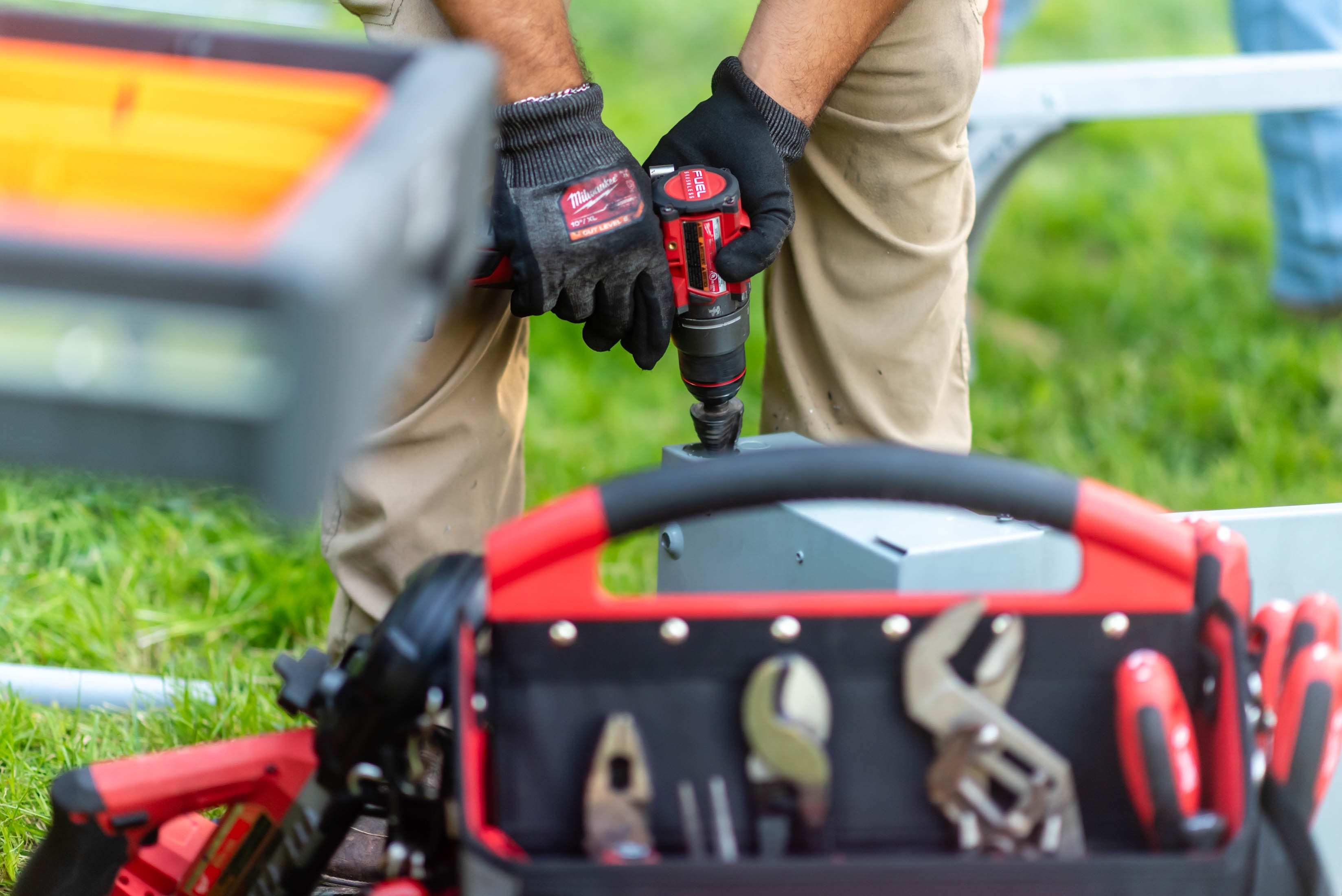 Gloved hands drilling into disconnect enclosure on solar racking with Milwaukee tool bag in foreground