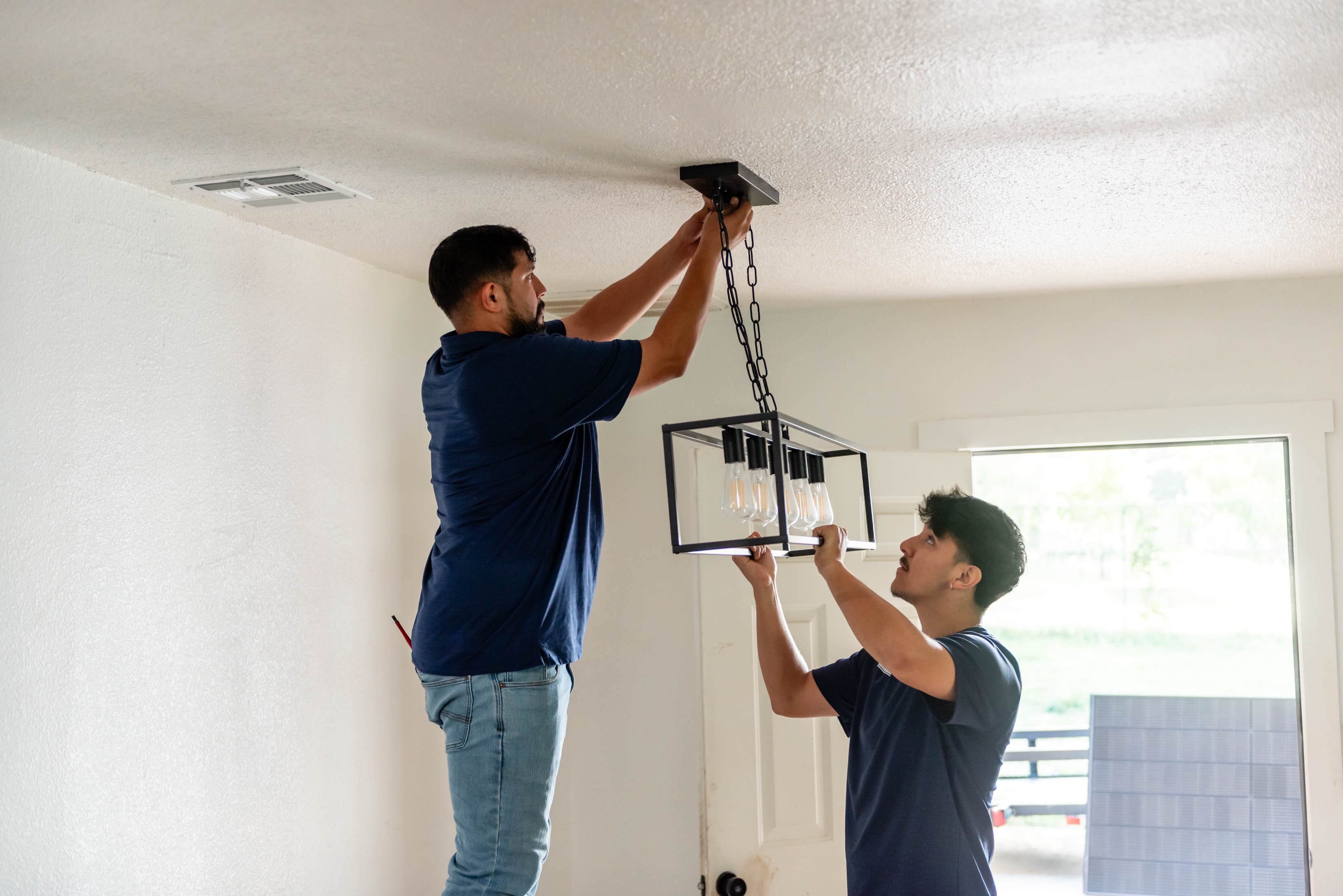 Two Casta Energy techs in navy polos installing black cage chandelier in residential dining room