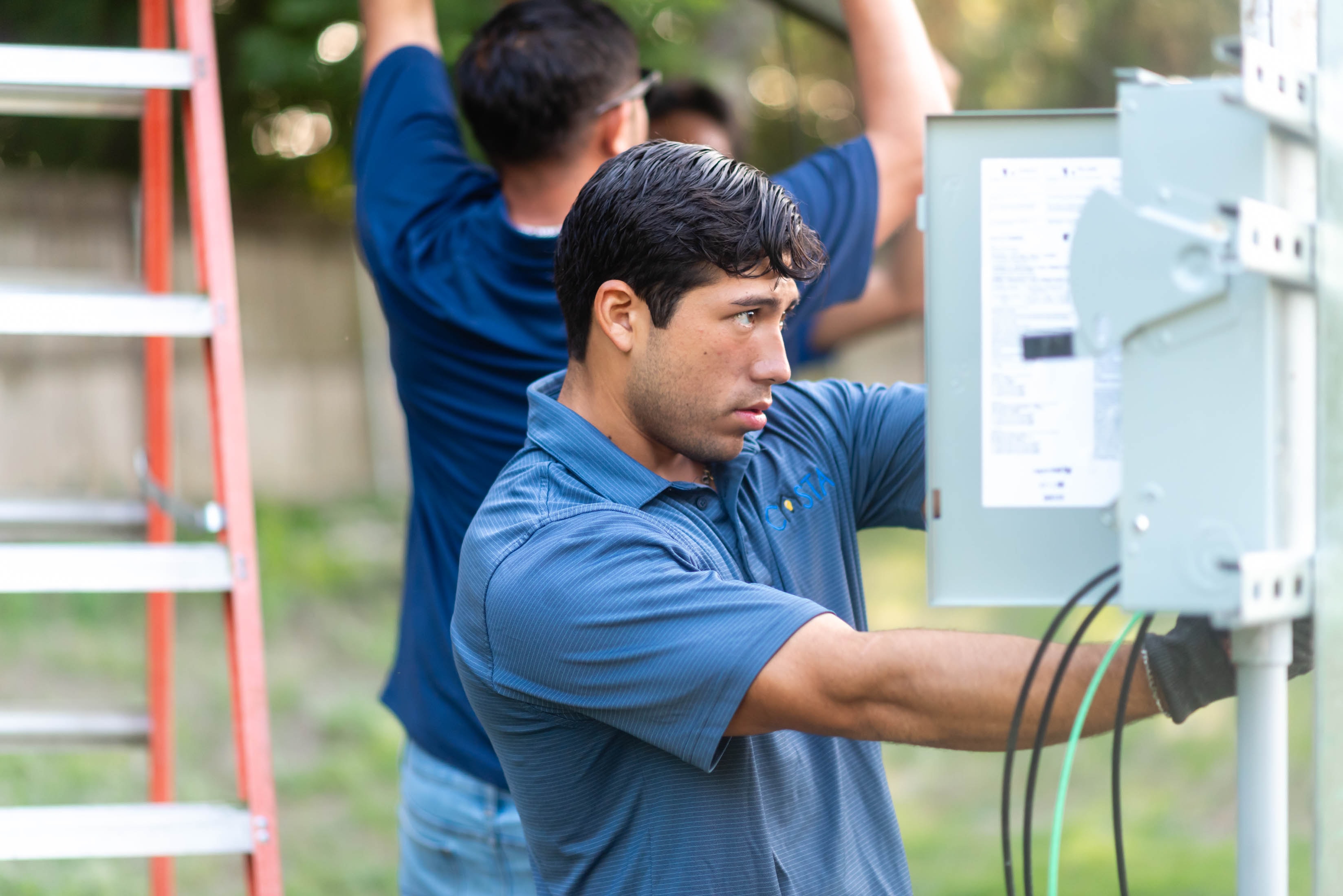 Two Casta Energy techs in blue polos wiring outdoor disconnect panel on solar racking — ground-mount install