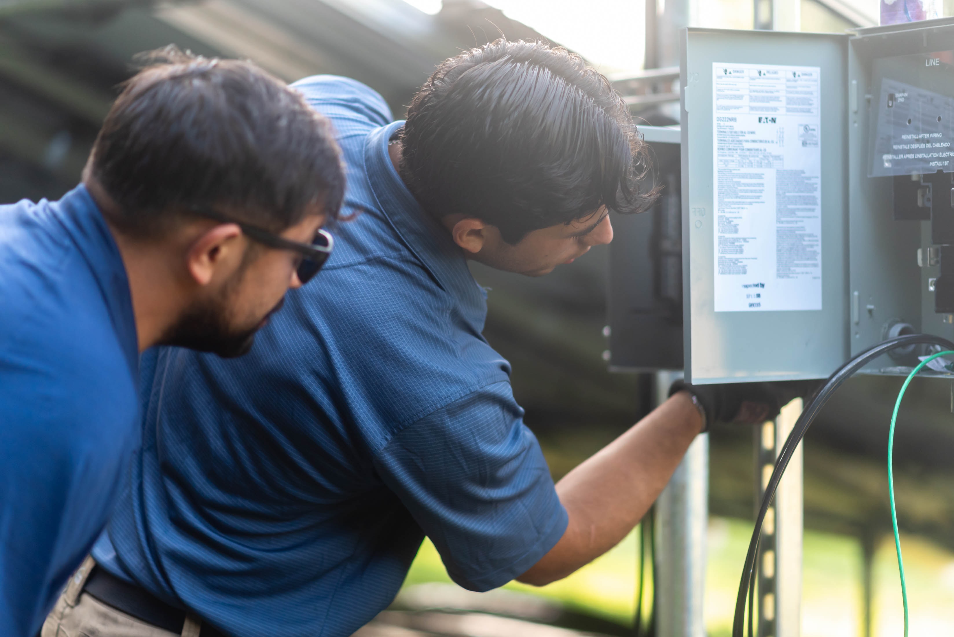 Two Casta Energy techs leaning in to wire Eaton disconnect panel on ground-mount solar racking